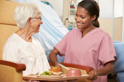 nurse serving a meal to a senior female patient who's sitting in a chair smiling